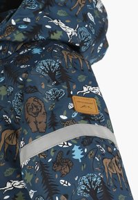 Lasten takki metsäeläin- ja puukuosisella sinisissä, ruskeissa ja vihreissä sävyissä, jossa on heijastinauha ja Swedemount-logo-ompelus.