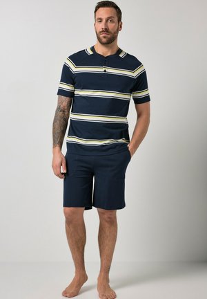 Man met een getatoeëerde arm, die een marineblauw gestreept kort mouw shirt en bijpassende korte broek draagt, blootsvoets staand tegen een effen achtergrond.