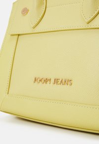 Žlutá texturovaná kabelka s prošívanými akcenty, která má zlatě embosované logo "JOOP! JEANS" a kulaté kovové detaily.