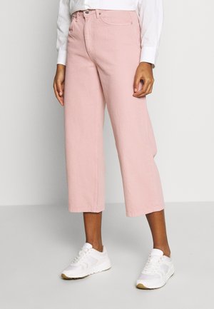 Persoon draagt een hoge taille, pastelroze, wijde cropped broek met witte sneakers en een witte longsleeve top, staand op een grijze vloer.