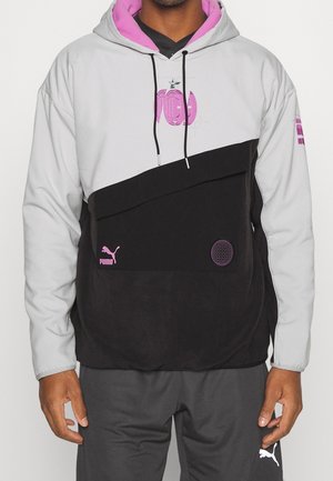 Mand iført en sort og grå hættetrøje med pink Puma-logoer og design, kombineret med mørkegrå Puma-bukser.