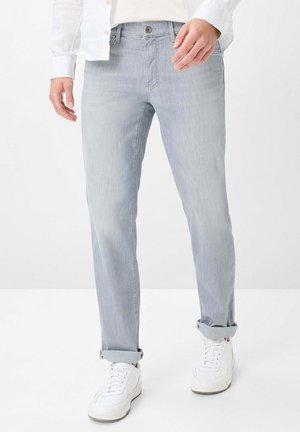Mann trägt hellgraue Jeans mit umgeschlagenen Säumen, weiße Sneakers und ein weißes Hemd mit Knöpfen, steht vor einem schlichten weißen Hintergrund.