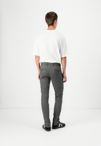Pantaloni grigi attillati con una finitura testurizzata, dotati di tasche posteriori e una toppa in pelle marrone, indossati con una maglietta bianca e sneakers nere.