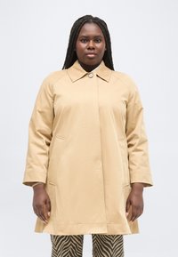 Γυναίκα με μακριά πλεξούδα, ντυμένη με μπεζ καμπαρντίνα πάνω από παντελόνι με ρίγες ζέβρας, στέκεται μπροστά σε ένα απλό φωτεινό φόντο.