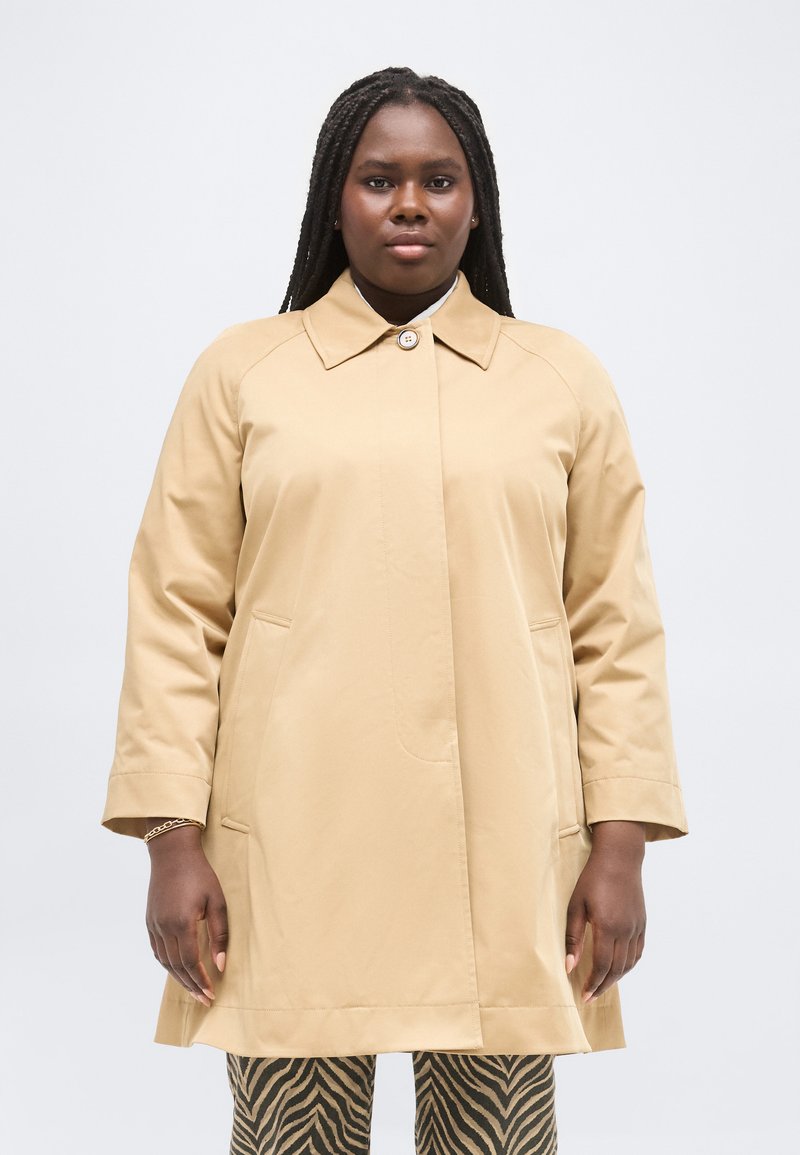 Γυναίκα με μακριά πλεξούδα, ντυμένη με μπεζ καμπαρντίνα πάνω από παντελόνι με ρίγες ζέβρας, στέκεται μπροστά σε ένα απλό φωτεινό φόντο.