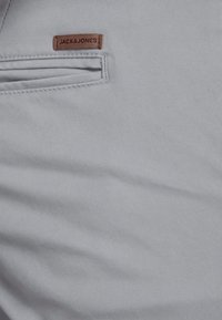 Szary materiał o gładkiej fakturze z małą brązową skórzaną metką z logo "JACK & JONES" w pobliżu szwu kieszeni.