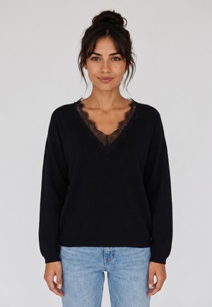 Donna con capelli scuri raccolti in uno chignon, indossa un maglione nero a V con bordo in pizzo e jeans azzurri, in piedi davanti a uno sfondo chiaro e semplice.