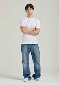 Maglietta bianca a maniche corte con logo nero, abbinata a jeans in denim azzurro chiaro e sneakers bianche, in piedi su uno sfondo neutro.