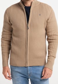 Cárdigan de cremallera beige con canalé y cuello alto, hecho de tejido texturizado. Presenta un pequeño logo y mangas ajustadas para un aspecto estilizado.