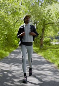 Pantaloni sportivi grigi con accenti neri, una giacca leggera grigia con zip e scarpe da ginnastica nere. La persona cammina su un sentiero fiancheggiato da alberi.