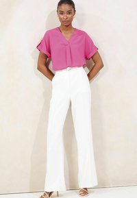 Blusa fucsia a maniche corte con scollatura a V infilata in pantaloni bianchi a gamba larga a vita alta; accentuata da una cintura chiara.