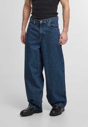 Wide-leg blue denim jeans with a high waist, featuring front pockets and a metal button closure, paired with black shoes.