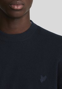 Suéter de punto azul marino con cuello redondo y un pequeño logo de pájaro bordado en el lado izquierdo del pecho. Textura suave, diseño casual.