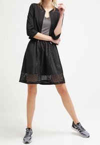 Czarna krótka kurtka z zamkiem błyskawicznym i rękawami długości trzy czwarte, w zestawieniu z perforowaną czarną spódnicą i szarymi trampkami.