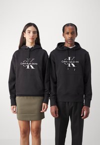 Crne dukserice s kapuljačom koje imaju prednje džepove i vezice, s bijelim logotipom "Calvin Klein Jeans" i grafičkim detaljima na prsima.