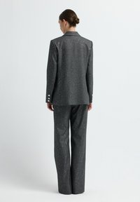 Frau, die sich abwendet, trägt einen dunkelgrauen Anzug mit dezenten silbernen Sprenkeln, ein Blazer mit Knöpfen an den Ärmeln und passende weit geschnittene Hosen.