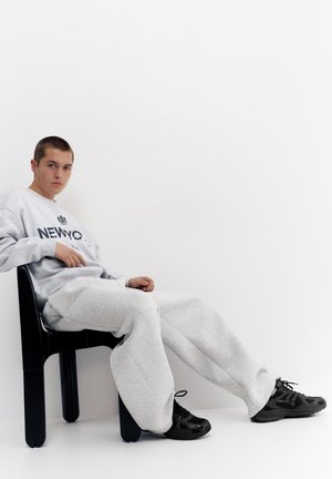 Jeune homme assis sur une chaise noire, portant un pantalon de jogging gris, un sweat-shirt avec le texte « NEW YORK » et des baskets noires sur un fond blanc.