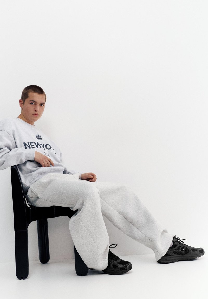 Jeune homme assis sur une chaise noire, portant un pantalon de jogging gris, un sweat-shirt avec le texte « NEW YORK » et des baskets noires sur un fond blanc.