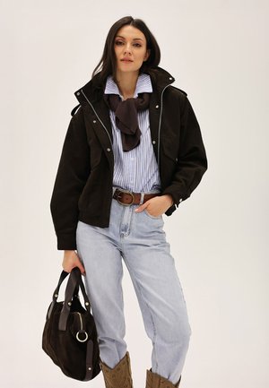 Femme portant une veste noire, une chemise rayée bleue, une écharpe marron, un jean clair, tenant un sac à main noir, debout devant un fond blanc uni.