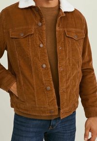 Braune Cordjacke mit einem weißen Fleece-Kragen, mit Knopfleiste, zwei Brusttaschen und einer strukturierten Oberfläche. Über einem braunen Pullover getragen.