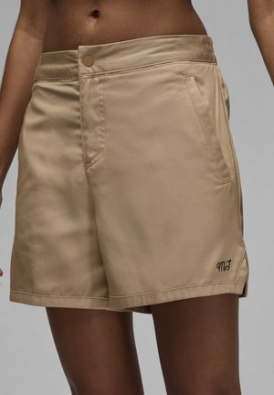 Hellbraune Shorts mit hohem Bund, Fronttaschen, Knopfverschluss und kleinen schwarzen bestickten Initialen am unteren linken Bein, getragen von einer Person.