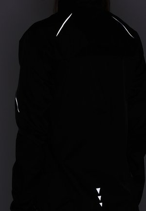 Person trägt eine schwarze Jacke mit reflektierenden weißen Streifen auf den Schultern, dem Oberarm und drei kleinen Dreiecken nahe dem unteren Rücken.