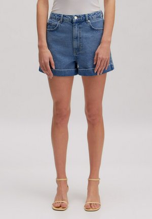 Frau trägt blaue, hochgekrempelte Jeansshorts, ein weißes Top und beige offene Sandalen mit Absatz, steht mit entspannten Händen neben dem Körper.