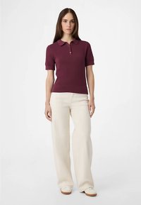 Bordeauxrotes Polohemd mit kurzen Ärmeln, Kragen, geripptem Stoff und Drei-Knopf-Leiste, kombiniert mit cremefarbenen Hosen mit weitem Bein und weißen Schuhen.