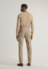 Suéter de punto beige combinado con pantalones de vestir beige claros que cuentan con bolsillos traseros y pestañas laterales ajustables, llevados con zapatos de ante marrón.