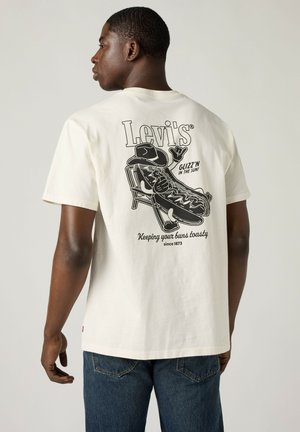 Mężczyzna w białym T-shircie Levi's z czarną grafiką hot doga odpoczywającego na krześle, z podpisem „Glizz'n in the sun! Trzymaj swoje bułeczki w cieple.”