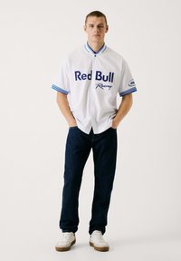 Witte kortarmige shirt met blauwe accenten en het logo "Red Bull Racing", gecombineerd met donkerdenim jeans en witte sneakers met bruine zolen.