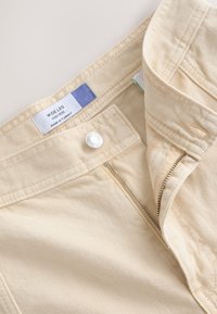 Pantalon beige à jambes larges et taille haute avec fermeture éclair et bouton. Tissu doux et texturé avec une étiquette indiquant "Fabriqué en Turquie".