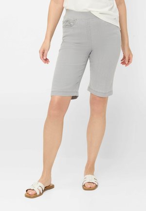 Femme portant un short gris clair à hauteur des genoux, un haut blanc et des sandales blanches faciles à enfiler, debout sur un fond blanc uni.