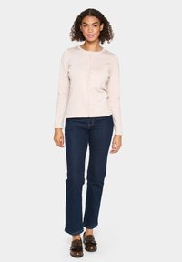 Cardigan long beige clair à manches longues avec un col rond et des boutons, associé à un jean bleu foncé et des mocassins à franges marron.