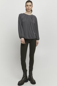 Grå langærmet bluse med plisseret halslinje, parret med mørkegrønne tætsiddende bukser og kraftige sorte ankelstøvler. Glat stof med afslappet pasform.