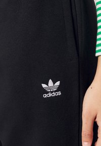 Gros plan sur un pantalon Adidas noir avec le logo trèfle blanc près de la poche latérale, une main posée à côté de la jambe, personne portant une manche rayée verte.