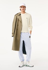 Beiger Pullover, olivgrüner Trenchcoat, hellblaue Jogginghose mit Karolining, und weiße Sneaker mit grünen Akzenten. Minimalistisches Design, lockere Passform.