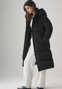 Cappotto lungo nero imbottito con cappuccio, dotato di chiusura con zip e tasche laterali, indossato sopra pantaloni grigio chiaro e sneakers. Texture liscia.