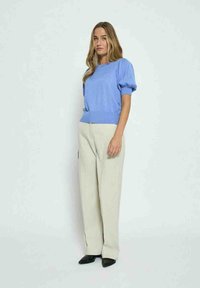 Soehe helesinine kudumüts lühikeste pallide varrukatega, koos kreemjate laia jala pükstega ja mustade terava ninaga kingadega, lihtsa tausta ees.