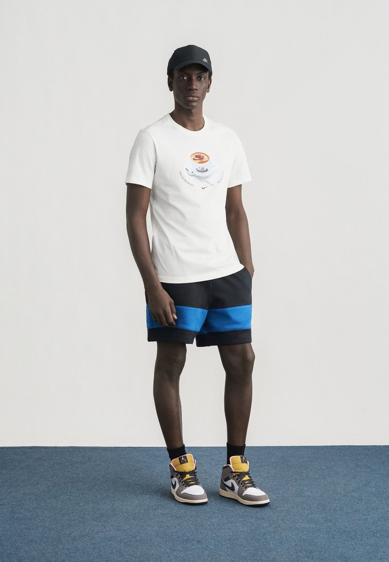 Giovane uomo che indossa una t-shirt bianca con grafica, pantaloncini neri e blu, cappellino nero e sneakers alte multicolore in piedi su un tappeto blu.