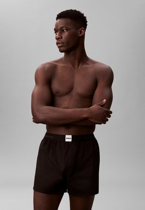 Schwarze Boxershorts mit einem Calvin Klein Logo am Bund. Das Modell posiert mit verschränkten Armen und präsentiert das Baumwollmaterial und die Passform.