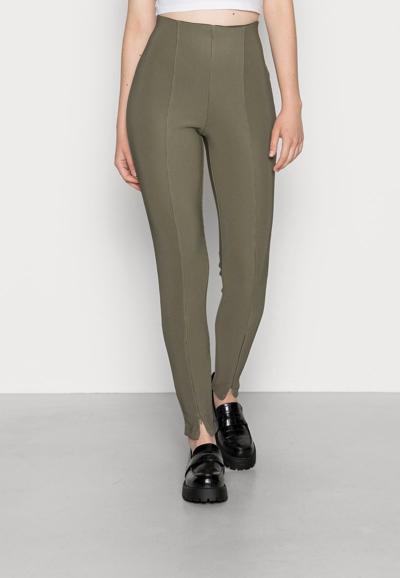 Kobieta w oliwkowych, prążkowanych legginsach z wysokim stanem, z przednimi szwami, oraz czarnych, masywnych loafersach, stoi na szarym podłożu przy białej ścianie.