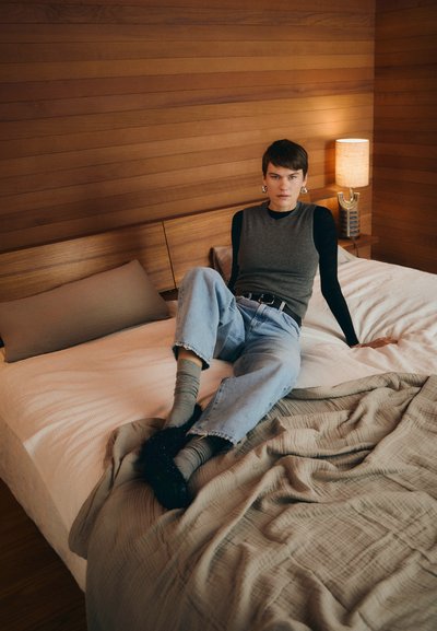 Grå ærmeløs top, sort langærmet skjorte, lyseblå denimjeans og teksturerede sokker. Neutralfarvet sengelinned og baggrund af trævæg.