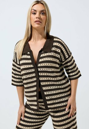 Donna con capelli biondi e lunghi che indossa un top a maglia a righe nere e beige con maniche corte e pantaloni abbinati su uno sfondo chiaro e uniforme.
