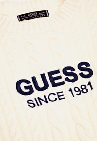 Pulover din tricot cu modele de cablu, de culoare crem, cu un text brodat proeminent în nuanță albastru marin: "GUESS" și "SINCE 1981". Guler ribatat, textură moale.