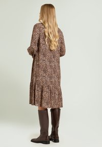 Leopardenmuster Midikleid mit Puffärmeln und gerafftem Saum, kombiniert mit braunen kniehohen Lederstiefeln. Minimalistischer grüner Hintergrund.