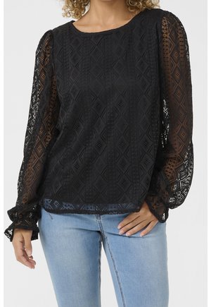 Kobieta w czarnej koronkowej bluzce z długim rękawem o geometrycznych wzorach oraz jasnoniebieskich dżinsach, z ręką opartą na biodrze.