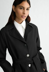 Cappotto nero con cintura, colletto a scollo con notched, bottoni argentati e una texture di tessuto liscia, indossato sopra una camicia bianca con colletto.