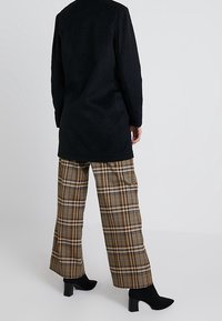 Schwarzer Fleece-Langmantel, beige und braun karierte weit geschnittene Hose sowie schwarze Knöchelschuhe, die kontrastierende Texturen und Muster zeigen.