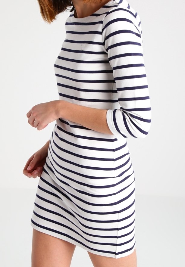 Femme portant une robe blanche avec des rayures horizontales bleu marine et des manches trois-quarts, montrée de la mi-cuisse au cou sur un fond uni.